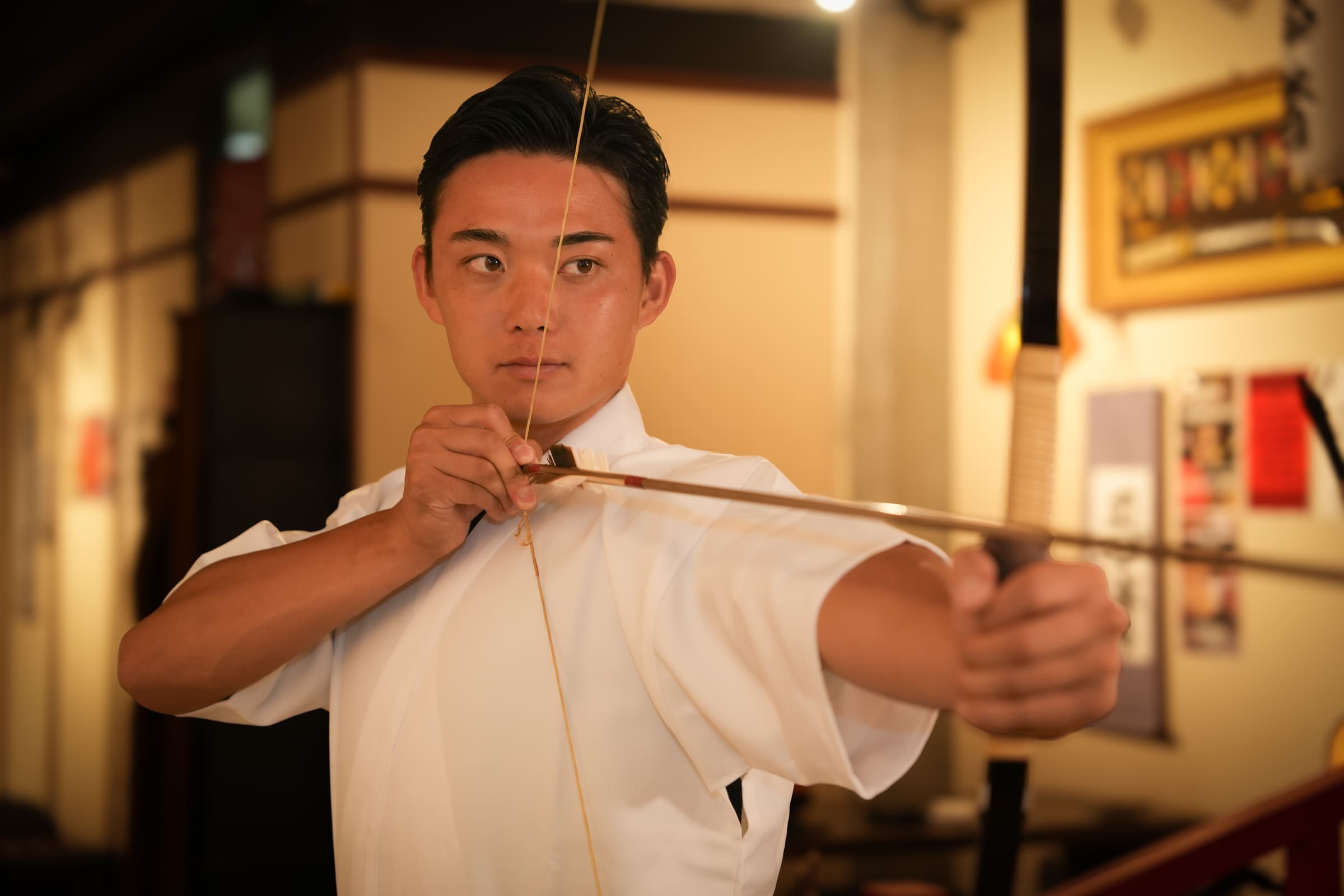 Man in white keikogi drawing a Japanese bow at full draw during an indoor kyudo archery experience