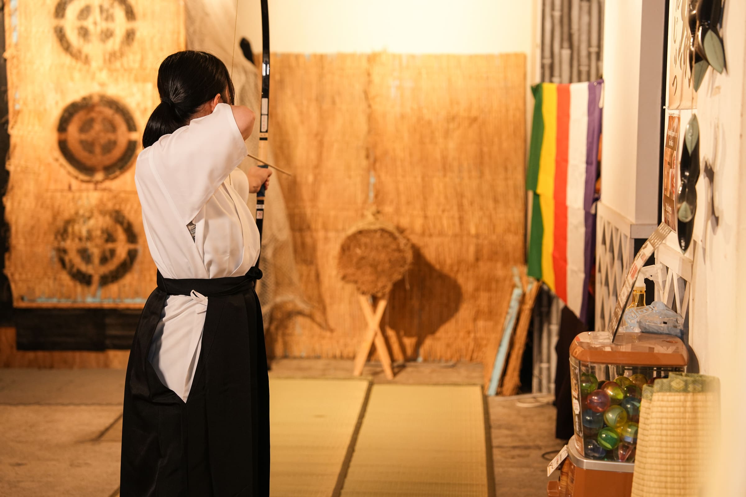 Archer in black hakama seen from behind aiming a kyudo bow at a straw target in a warmly lit Japanese archery studio