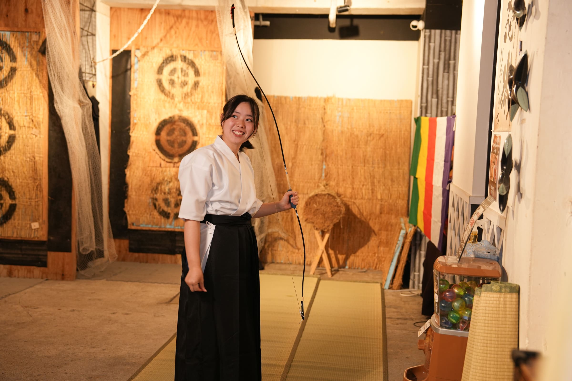 Smiling guest in hakama holding a traditional kyudo bow inside a Japanese archery experience space with straw targets