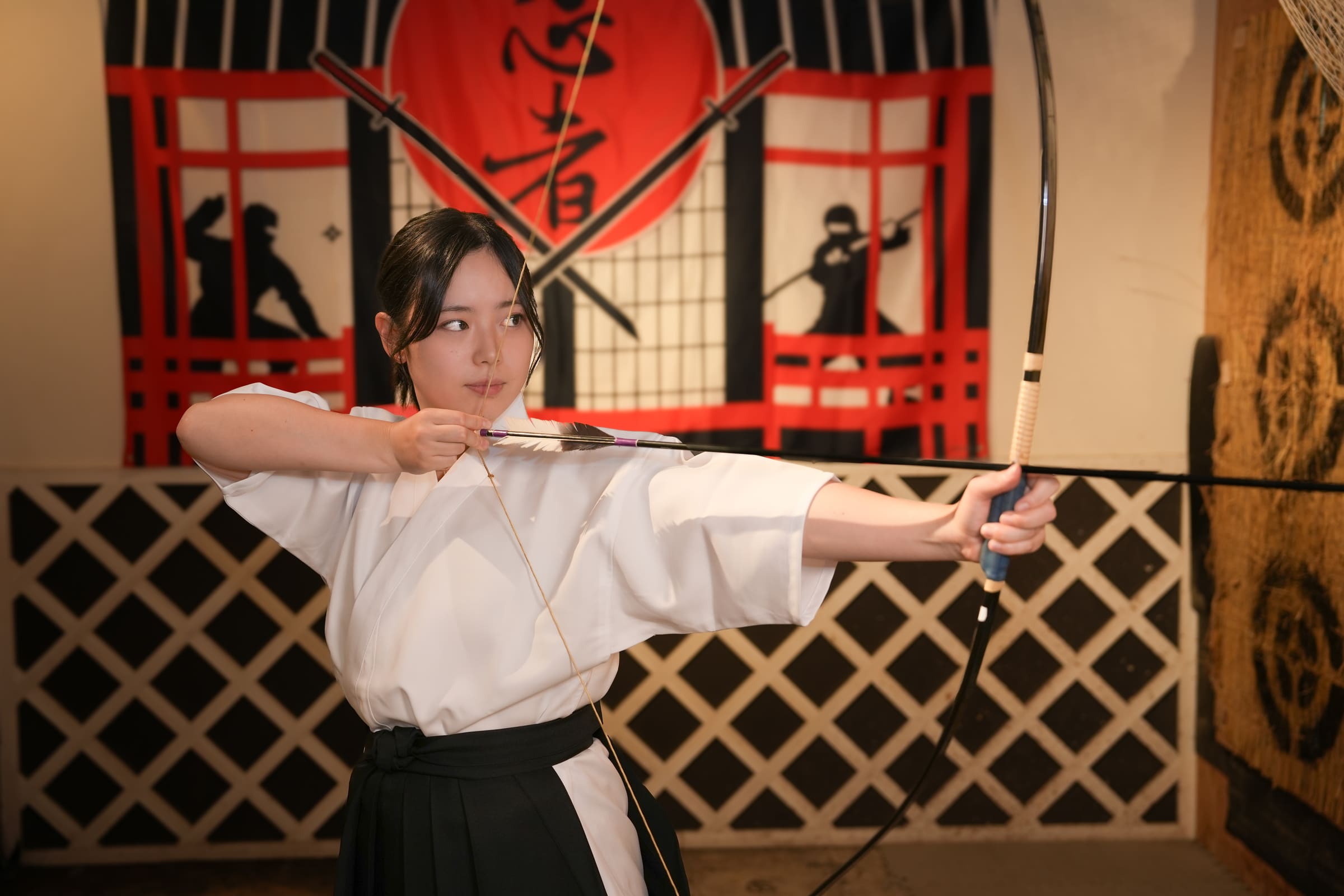 Focused archer in white top and black hakama drawing a Japanese bow during a kyudo experience with ninja decor behind