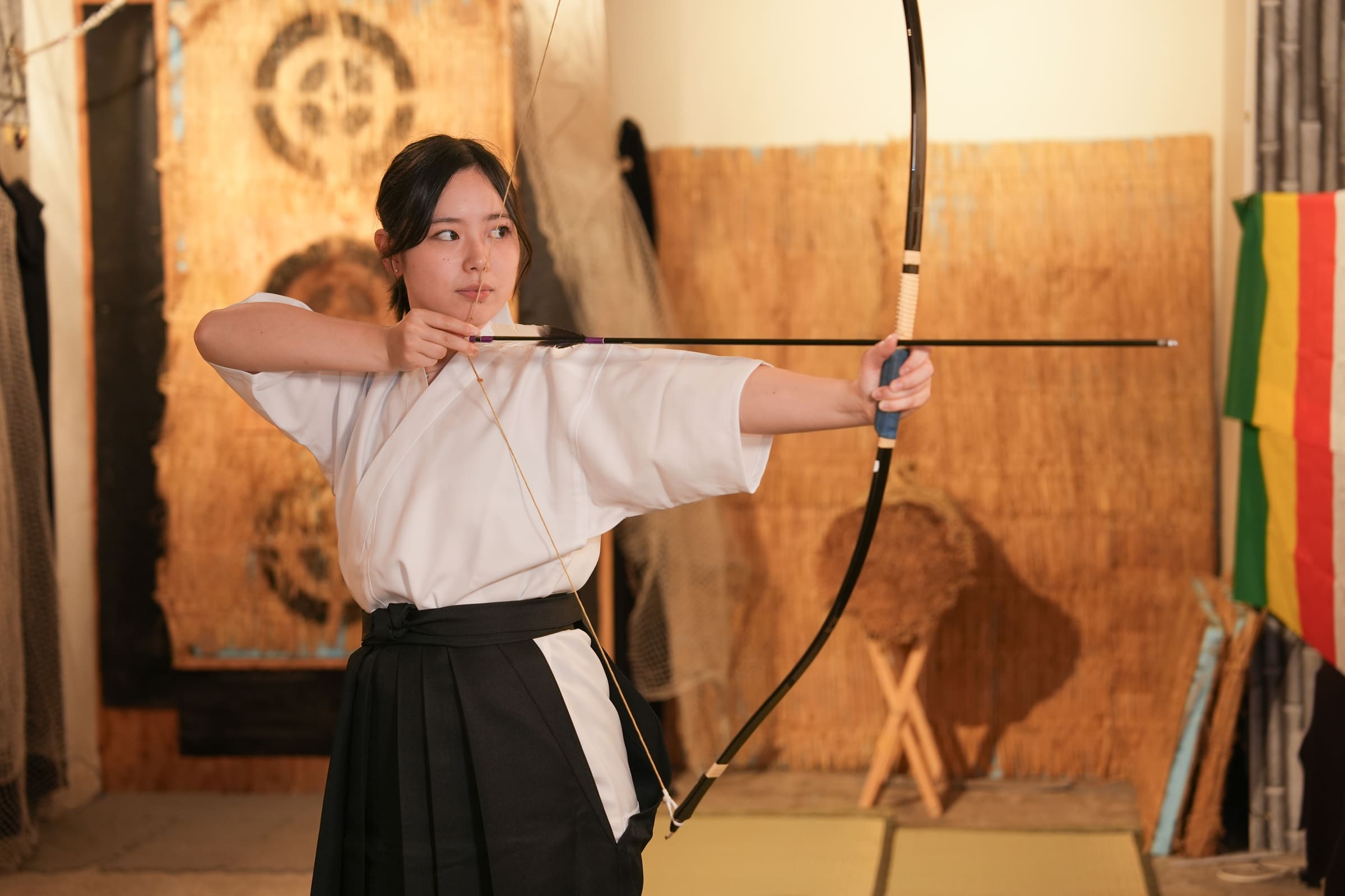 Archer in black hakama at full draw with a kyudo bow beside a straw makiwara target in a warmly lit Japanese archery room