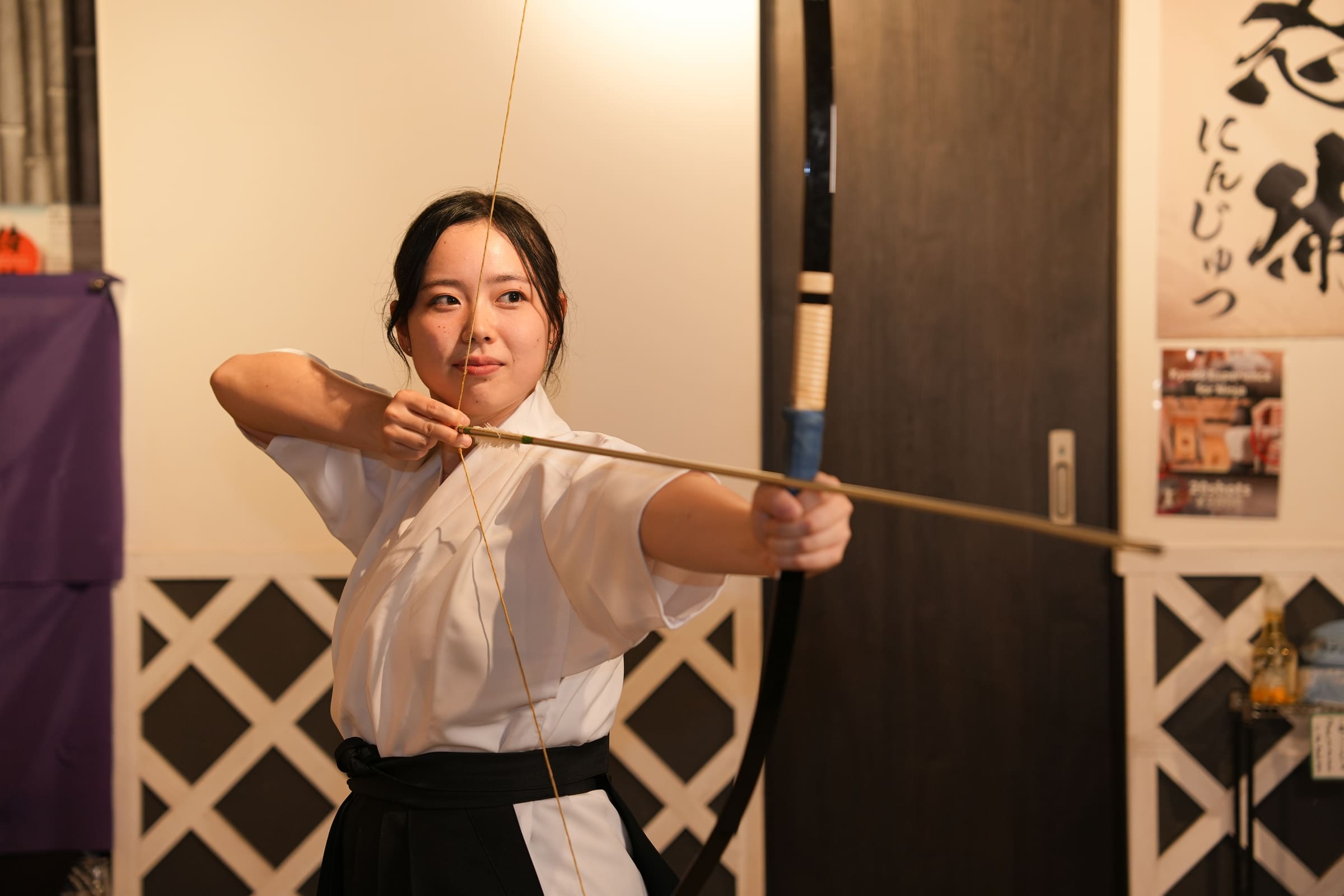 Woman in white keikogi and black hakama drawing a Japanese kyudo bow at full draw during an indoor archery experience