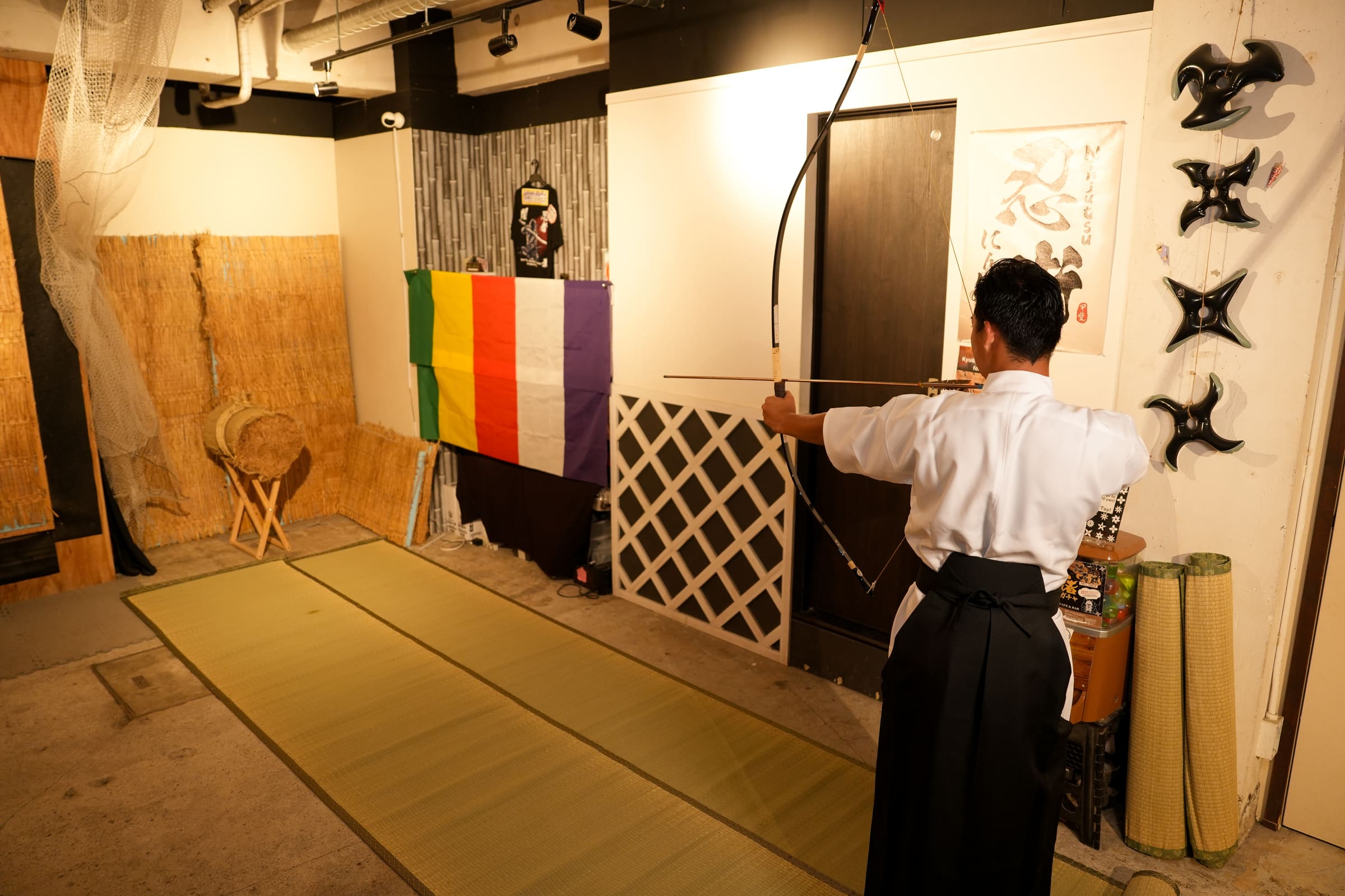 Indoor kyudo experience space with tatami mats and straw target, a guest in hakama aiming a Japanese bow