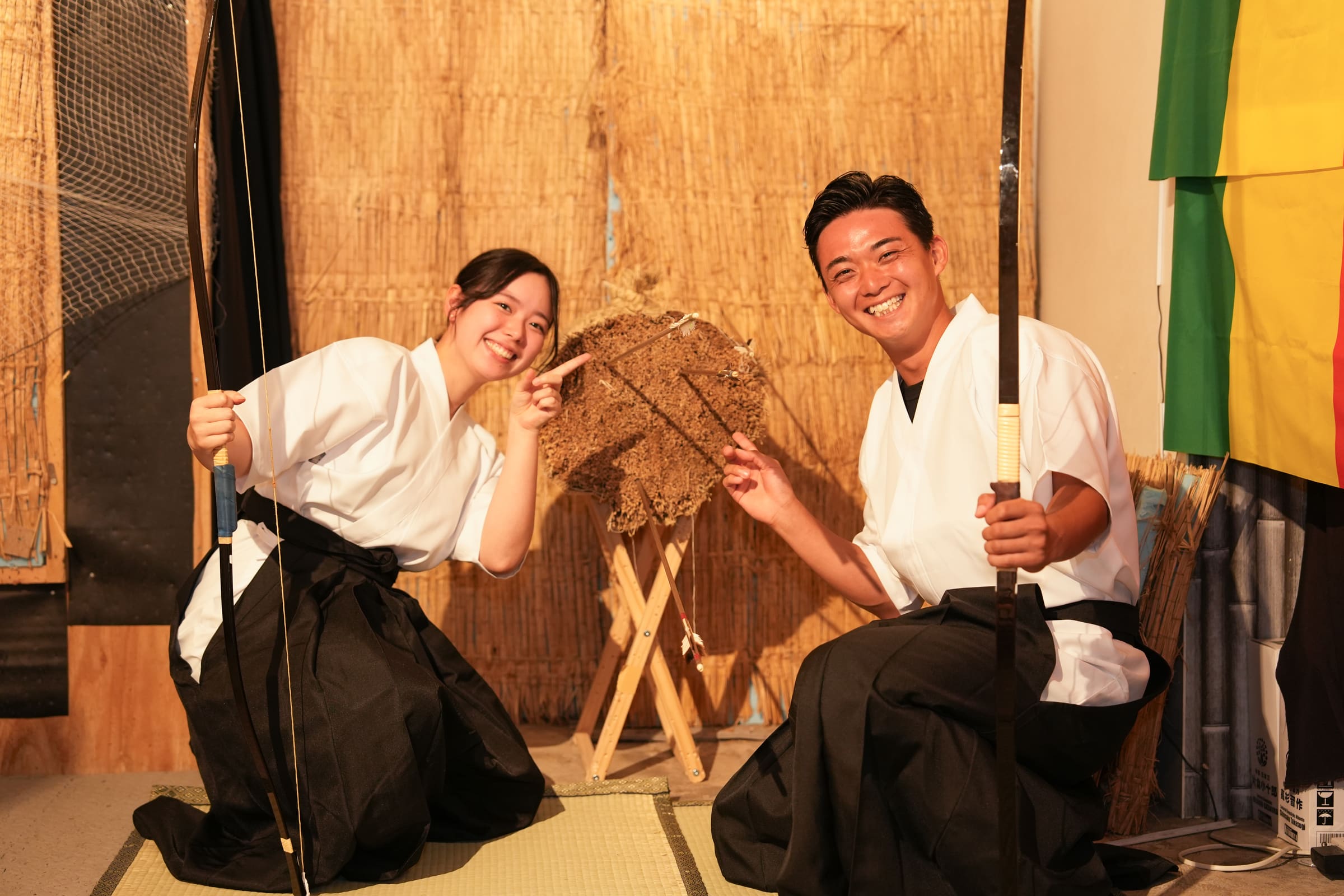 Friends in white keikogi and black hakama kneeling on tatami with bows, celebrating hits on a straw kyudo target