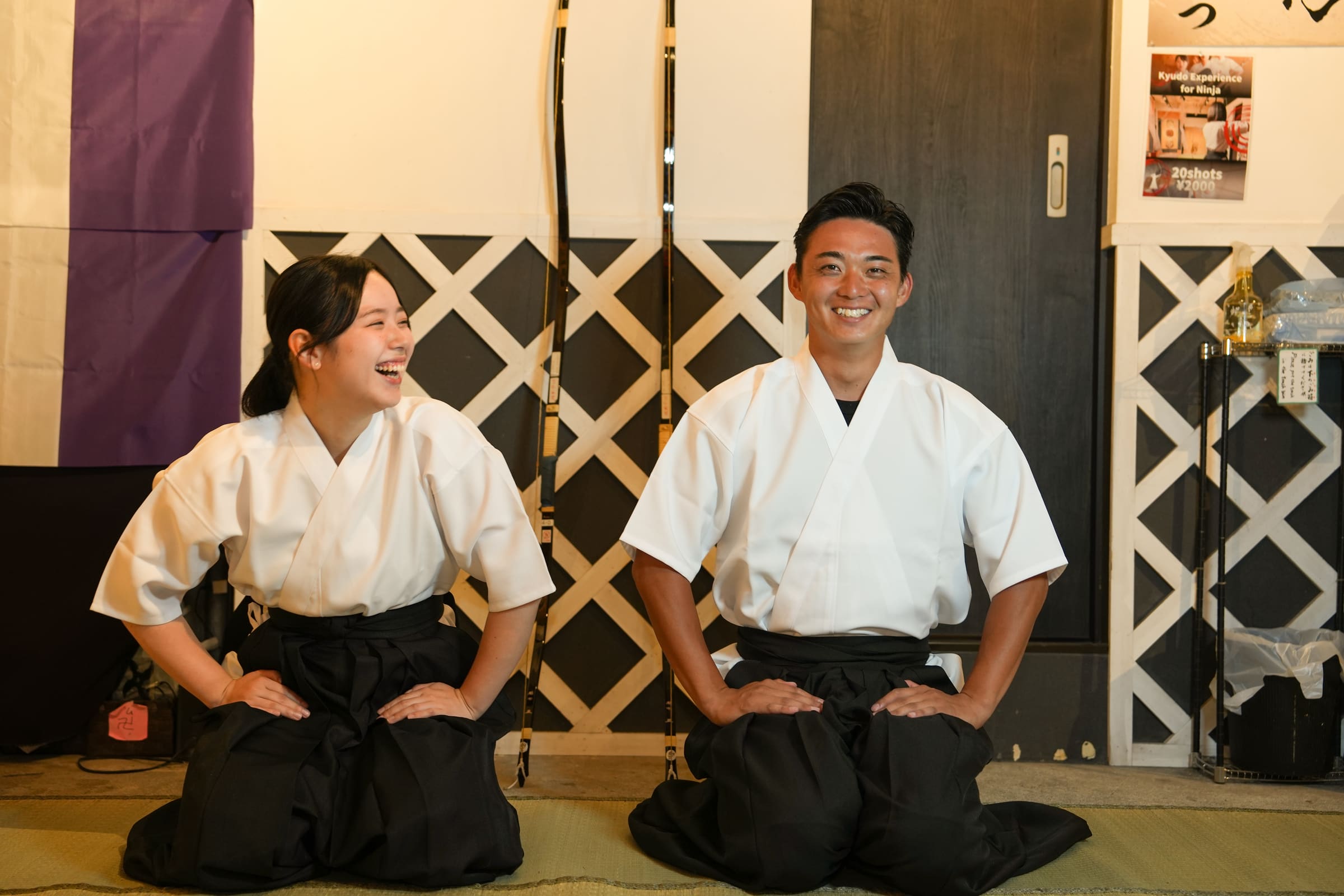 Two guests in kyudo uniforms with black hakama sitting seiza on tatami and laughing during a Japanese archery experience