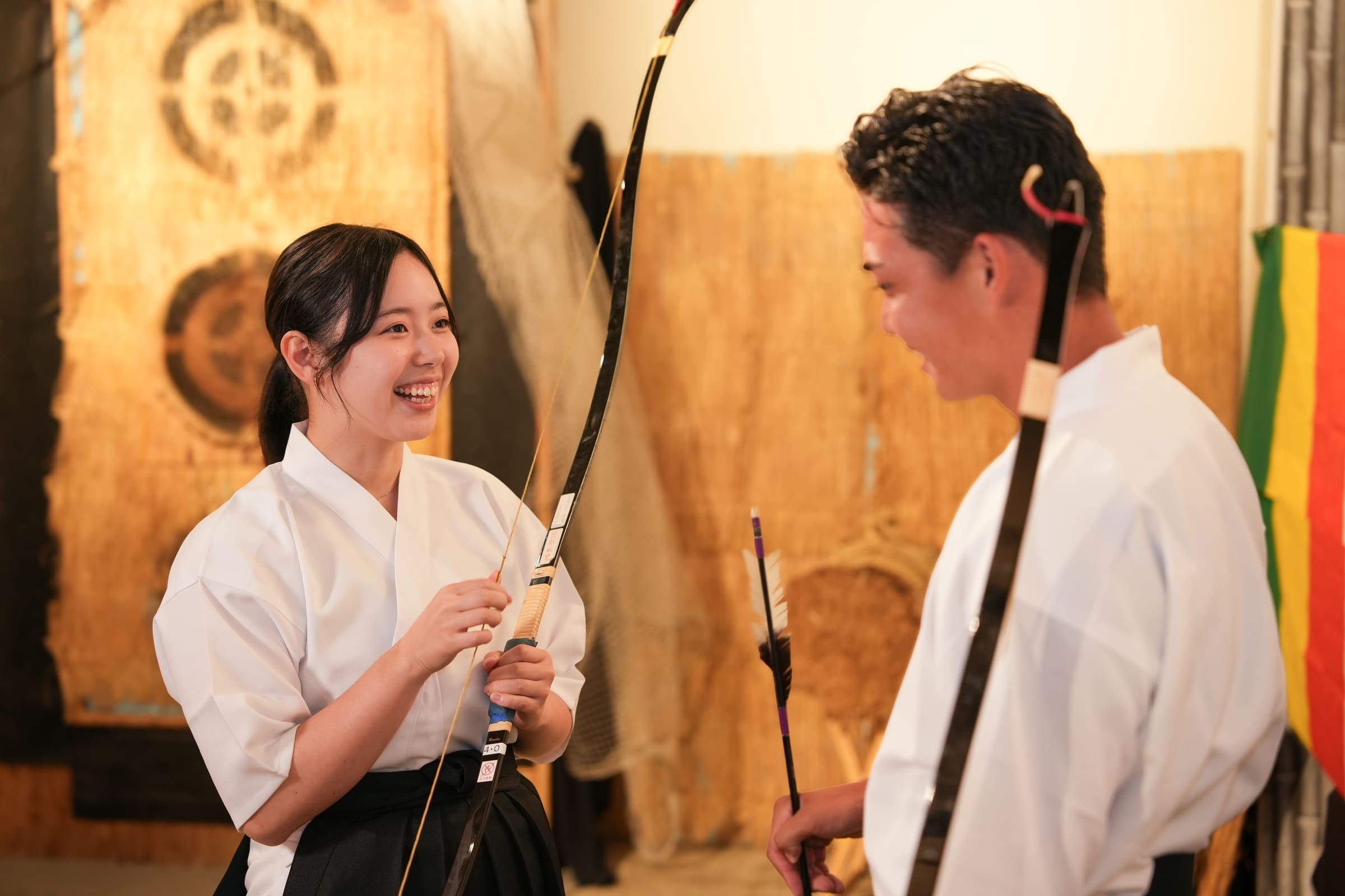 Woman in hakama smiling brightly with a Japanese bow during a kyudo archery experience with a friend