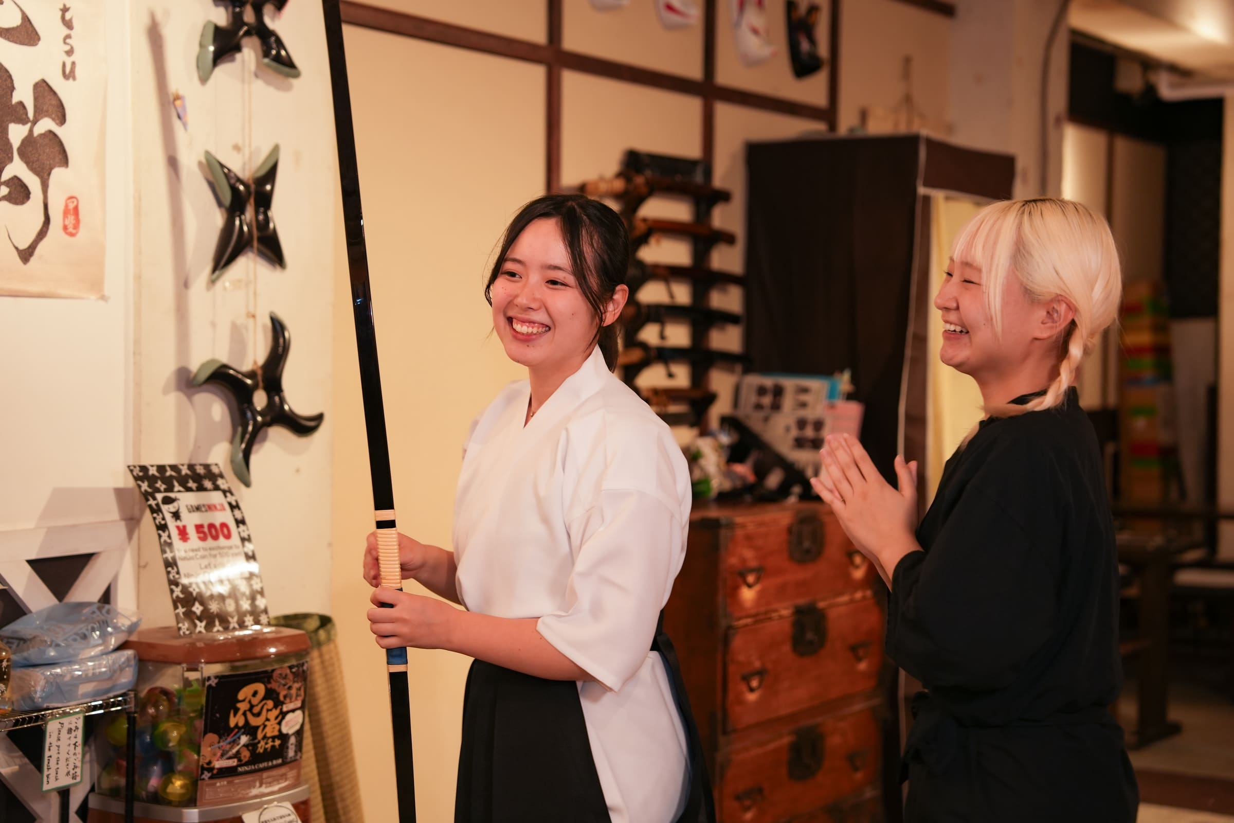 Guest in keikogi and hakama laughing while holding a Japanese kyudo bow in a warmly lit experience cafe