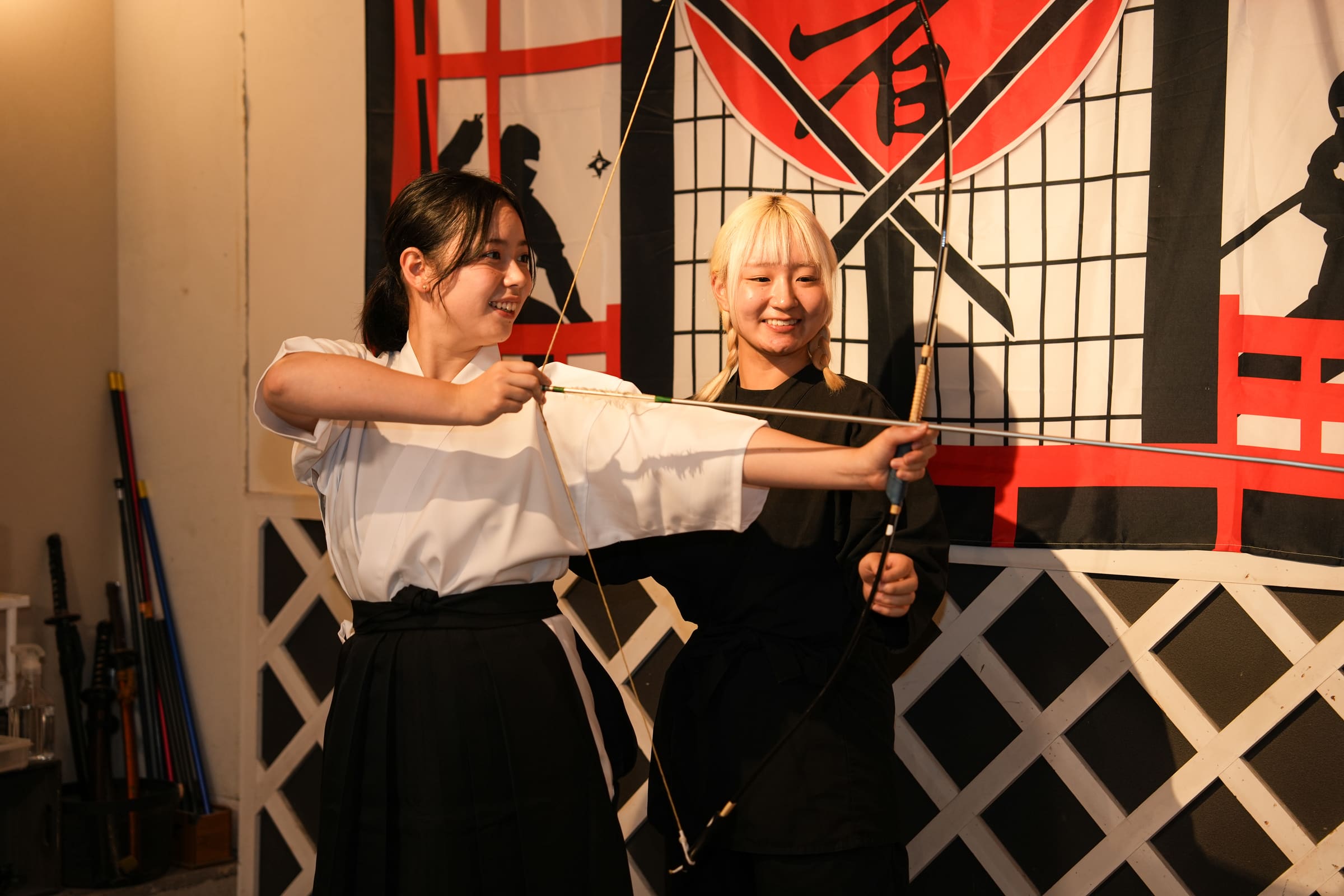 Guest in hakama aiming a kyudo bow with her instructor beside her, framed by red and black Japanese banners