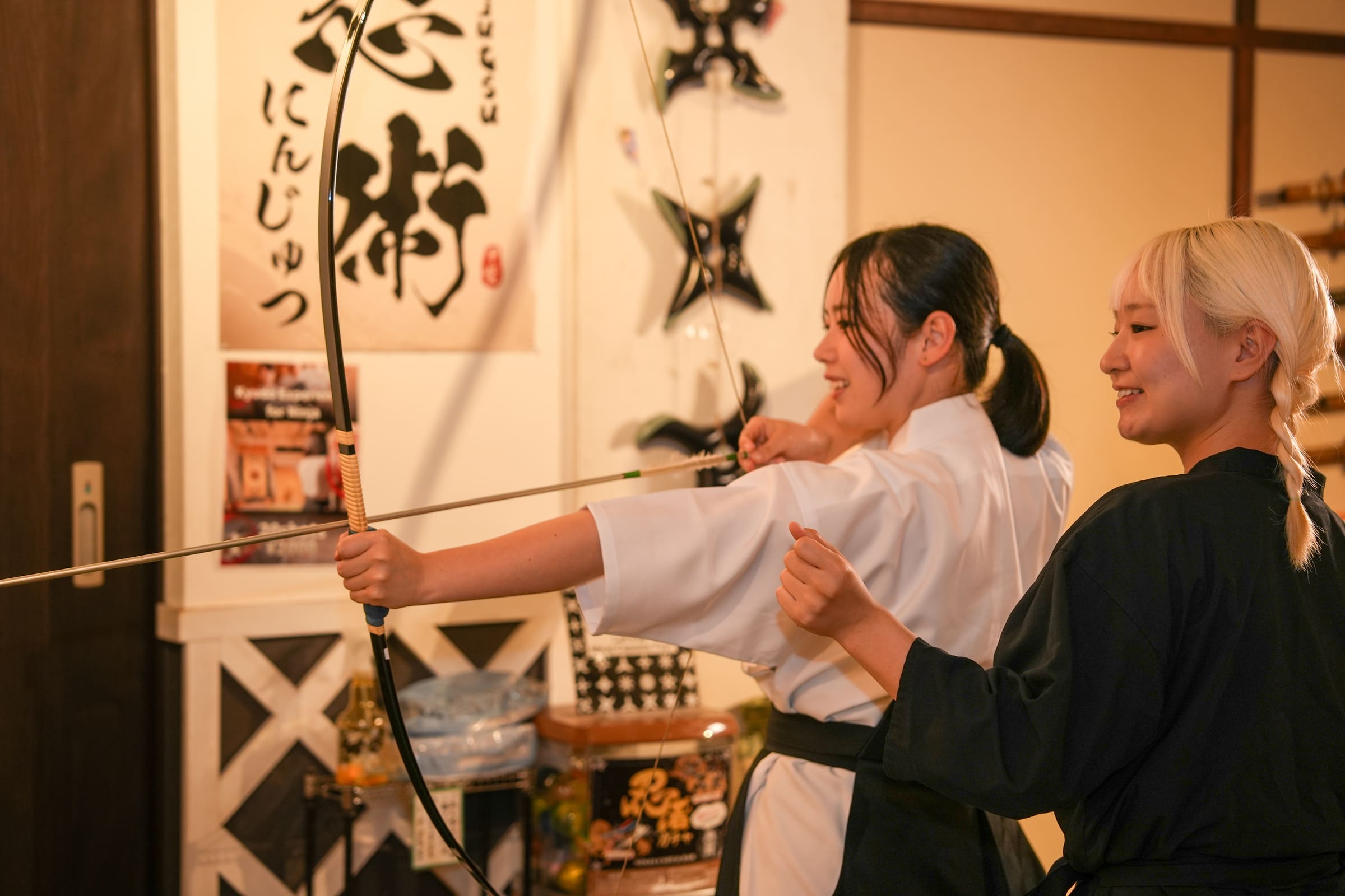 Guest in white keikogi and hakama at full draw of a Japanese kyudo bow during an indoor archery experience