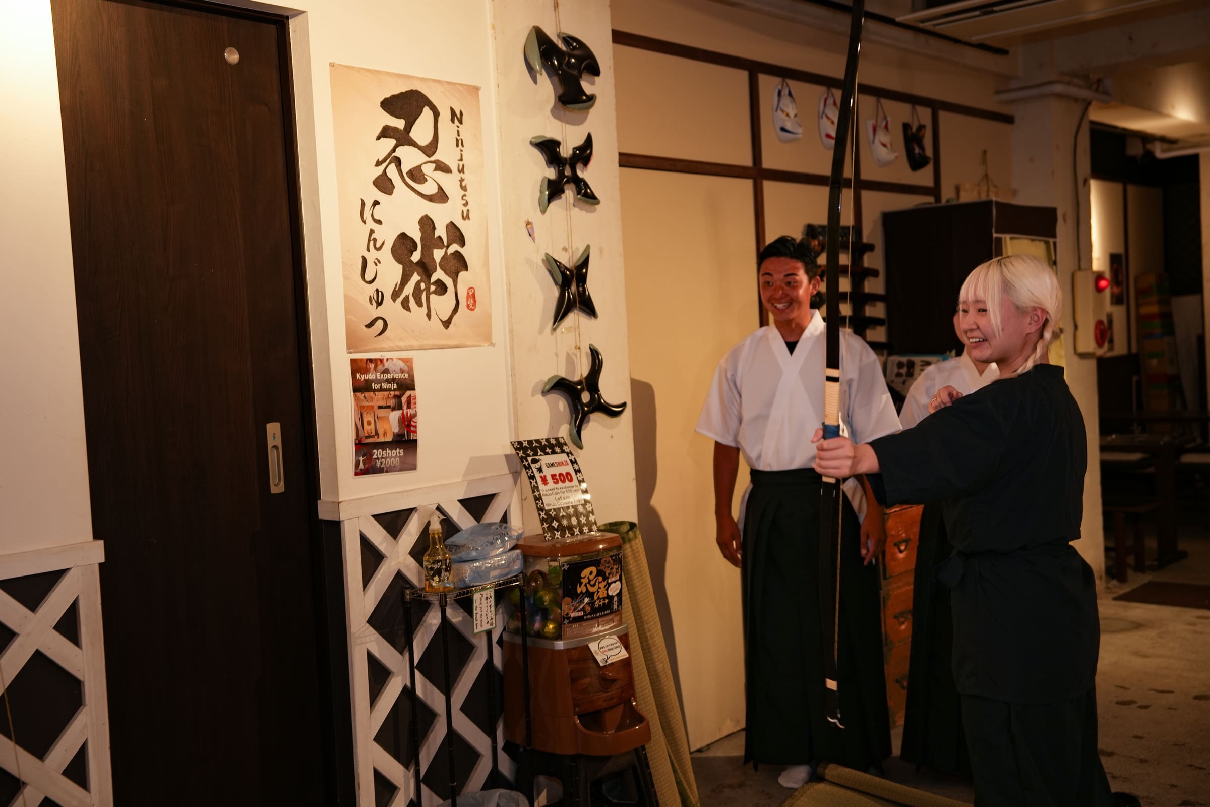 Guest raising a Japanese yumi bow beside an instructor in hakama at a kyudo experience inside a ninja-themed cafe